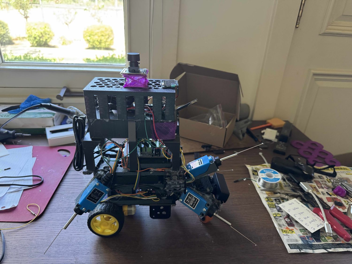 Low front close-up at table level, showing two orange castor wheels, a black webcam module, and spring-steel whisker feelers splayed left and right.