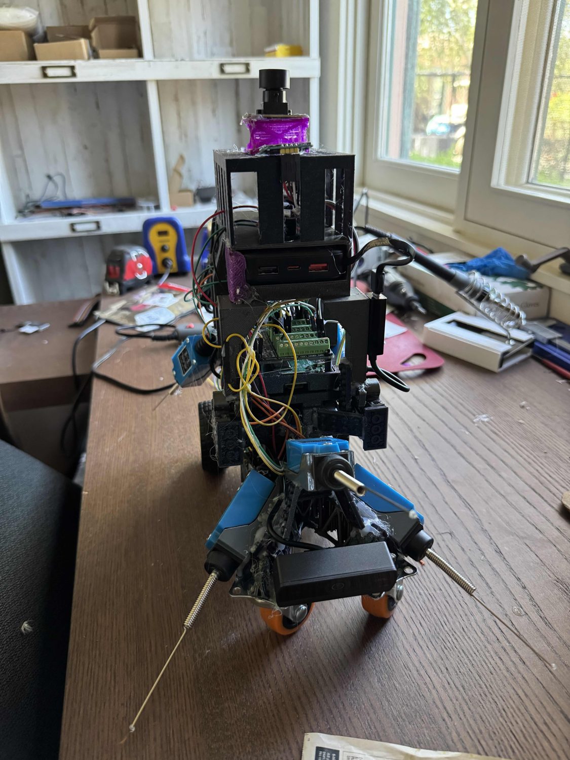 Front view of the R3M1 robot, full height, showing the camera on top, the printed cage, the Raspberry Pi, a green screw-terminal board, and orange castor wheels at the base.